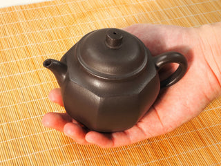 Hand holding Shihuang Bafang Yixing teapot in palm over bamboo mat, showing size and dark faceted octagonal body