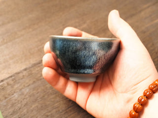 Handheld side view of a Shu Kou frosted hundred-flower Jianzhan bowl, showing the classic contracted mouth, soft matte blue-grey crystalline patches across the dark exterior, and a small unglazed foot.