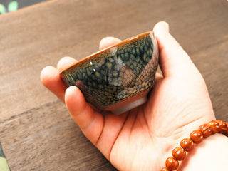 Handheld side view of a Shu Kou green partridge Jianzhan bowl, showing the classic contracted mouth, dense olive-green partridge speckles on the exterior, and an orange-gold rim.