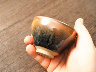 Handheld side view of a Shu Kou hare's-fur Jianzhan bowl, showing the classic contracted mouth, dense dark brown-to-black streaks on the exterior, and a small unglazed foot.