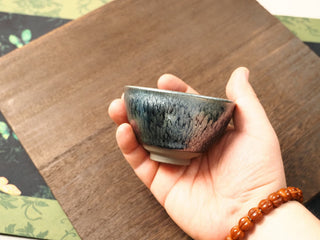 Handheld side view of a Shu Kou hundred-flower Jianzhan bowl, showing the classic contracted mouth, dense crystalline pattern in dark blue-black with a soft iridescent sheen on the exterior, and a small unglazed foot.