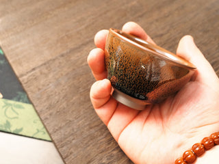 Handheld side view of a Shu Kou yellow partridge Jianzhan bowl, showing the classic contracted mouth, rust-orange speckles on the exterior, and a small unglazed foot.