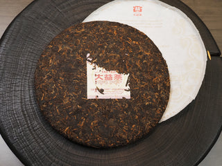 Unwrapped 2019 Dayi 7572 ripe puerh tea cake showing a compressed dark brown leaf surface with the embedded trademark paper (Neifei) on a wooden board.
