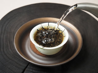 Hot water being poured over Yunnan Shaken-Leaf Black Tea in a white porcelain tasting cup.