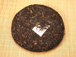 Front of Jing Chang Hao Mengsong Autumn Harvest 2013 raw Pu-erh 357g cake, loose arbor leaves with white nei fei label on a bamboo mat