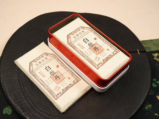 Tin box open showing one 2014 Fuding White Peony waffle cake inside, with another wrapped cake displayed in front.
