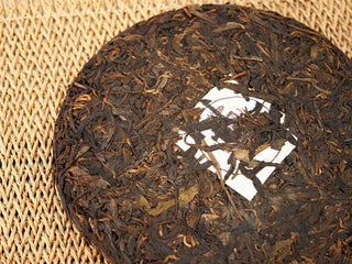 Close-up of Jing Chang Hao Quiet Years 2018 raw Pu-erh cake surface, glossy dark-brown arbor leaves pressed around the white nei fei label