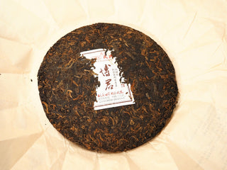 Unwrapped 2020 Bojun Ripe Pu-erh 500g cake from above on rice paper, dark fermented leaves with golden buds and inner ticket.