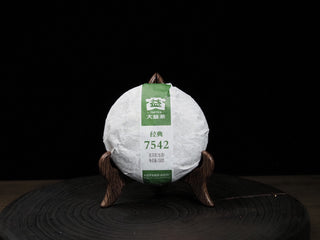 A 150g cake of 2020 Dayi 7542 raw puerh tea wrapped in trademark paper and placed on a wooden stand.