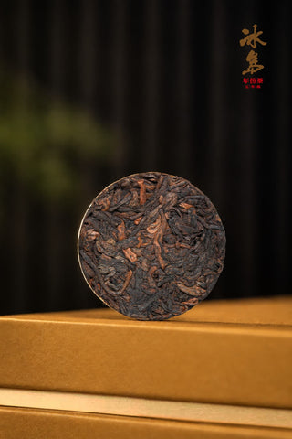 Single 8g coin cake of Mengku Rongshi Bingdao Aged Ripe Pu-erh, dark fermented leaves with golden tips, on the gold gift box.
