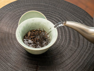 Pouring hot water from a kettle into a celadon gaiwan to brew Mang Fei ripe pu-erh tea.