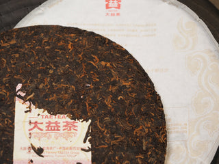 Close-up of 2019 Dayi 7572 ripe puerh tea cake surface, showing the embedded inner trademark ticket (Neifei) alongside the large brand description paper (Neipiao).