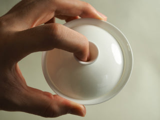 Hand holding pure white egg-shell porcelain gaiwan lid, finger inside the lid knob, glossy white glaze