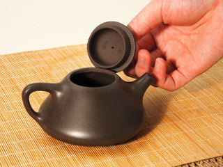 Hand lifting the lid of a Fengming Ziye Shipiao Yixing teapot, showing the lid underside seals and the open pot mouth