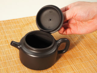 Hand lifting the lid of a Heini Dezhong Yixing teapot, showing the lid underside and open pot mouth on bamboo mat
