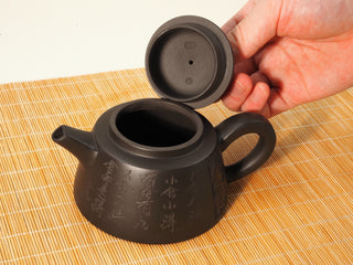 Hand lifting the lid of a Black Zhu Ni Shede Yixing teapot, showing the lid underside seal and open body on bamboo mat