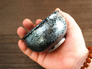 Another handheld side angle of a Jiang Jun Bei Jianzhan tea bowl, displaying the rounded form and metallic silver oil-spot crystals on the exterior.