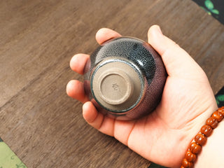 Base of a Lan Qi Lin Shu Kou Jianzhan bowl held in hand, showing the unglazed foot ring, a small impressed makers mark, and the exterior thinning to a russet tone at the foot.