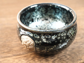 Side view of a Rui Shou sky eye oil spot Jianzhan bowl on a wooden table, showing the rounded silhouette and a raised clay medallion on the body.