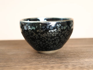 Front view of a Wu Zu Bo sky eye oil spot Jianzhan tea bowl on a wooden table, showing the deep open form and dense silver-ring oil-spot crystals on the exterior.