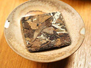 2018 sun-dried Fuding White Peony compressed tea biscuit in a small ceramic dish, top view showing the leaf texture and pressed square shape.