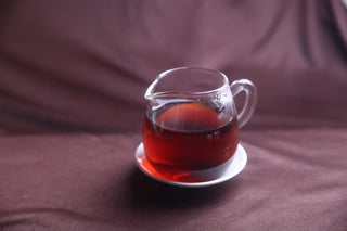 Clear and bright reddish-brown infusion of 2017 aged ripe puerh tea in a glass fair cup.