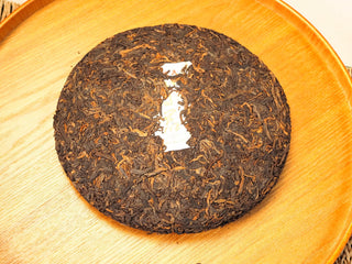 Unwrapped Bojun Ripe Pu-erh 7.0 (2026) 500g cake from above, dark fermented leaves with golden buds on a wooden tray.