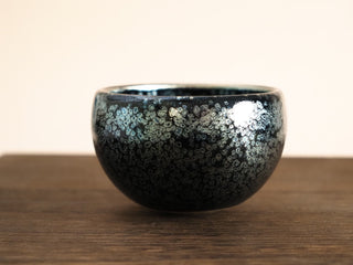 Front view of a Bu Dao Weng sky eye oil spot Jianzhan tea bowl on a wooden table, showing the round-bellied form and a wide unglazed foot.