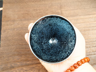 Handheld top-down view of a Da Dou Li sky eye oil spot Jianzhan bowl in palm, showing densely packed silver oil-spot crystals across the entire interior.