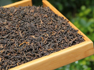 Close-up of dark, oily ripe puerh leaves and golden rose accents in a wooden tea tray.