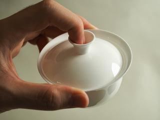Hand holding pure white egg-shell porcelain gaiwan with lid on, top-down view of glossy white glazed surface