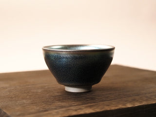 Front view of a Lan Qi Lin Shu Kou Jianzhan tea bowl on a wooden table, showing the slightly inward-curved rim, dense fine silver-blue pinpricks on the exterior, and a small foot.