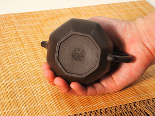Hand showing octagonal base of Shihuang Bafang Yixing teapot with round maker's seal stamp, bamboo mat background