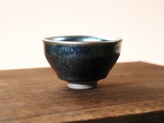 Front view of a Shu Kou frosted hundred-flower Jianzhan tea bowl on a wooden table, showing the contracted-mouth silhouette, soft matte dark blue-black exterior with quiet crystalline patches, and a small unglazed foot.