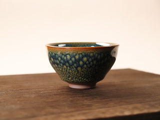 Front view of a Shu Kou green partridge Jianzhan tea bowl on a wooden table, showing the contracted-mouth silhouette, olive-green partridge speckles on the exterior, and a small unglazed foot.