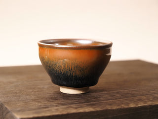 Front view of a Shu Kou hare's-fur Jianzhan tea bowl on a wooden table against a light background, showing the contracted-mouth silhouette, dark brown-to-black streaks on the exterior, and a small unglazed foot.