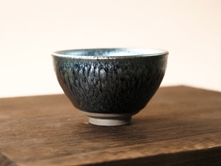 Front view of a Shu Kou hundred-flower Jianzhan tea bowl on a wooden table, showing the contracted-mouth silhouette, dark blue-black crystalline exterior with subtle iridescence, and a small unglazed foot.