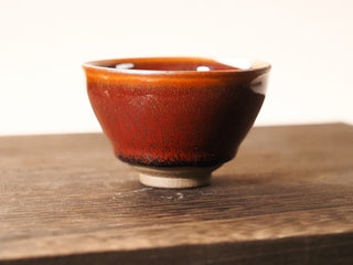 Front view of a Shu Kou red hare's-fur Jianzhan tea bowl on a wooden table against a light background, showing the contracted-mouth silhouette, dark streaks on the red-orange exterior, and a small unglazed foot.