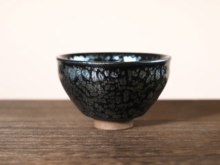 Front view of a Shu Kou sky eye oil spot Jianzhan tea bowl on a wooden table, showing the slightly inward-curved rim and dense silver exterior crystals.