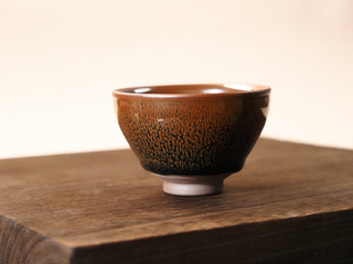 Front view of a Shu Kou yellow partridge Jianzhan tea bowl on a wooden table, showing the contracted mouth, rust-orange speckles across the exterior, and a small unglazed foot.