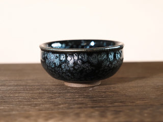 Front low-angle view of a Xiang Lu Jianzhan tea bowl with large silver oil-spot crystals and blue-green iridescence on the dark exterior, on a wooden surface.