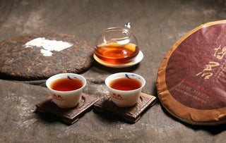 2017 Bojun Ripe Pu-erh brewing scene: wrapped cake, unwrapped cake, glass pitcher of red liquor, two small cups.