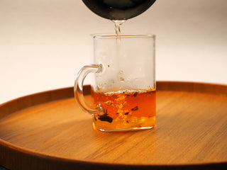 Hot water being poured into a clear glass mug, releasing the deep amber color of brewed 2018 sun-dried White Peony compressed tea biscuit.