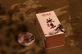 A single silver-wrapped Mengku Rongshi 2020 Bingdao Aged Raw Pu-erh coin cake beside its small gift box, on a brocade cloth.