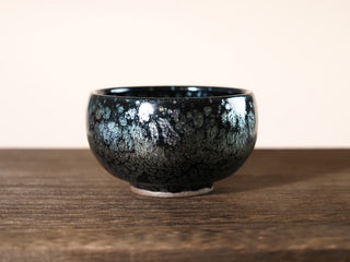 Front low-angle view of a Da Chan Ding Jianzhan tea bowl showing the short broad silhouette and silver oil-spot pattern on the dark exterior, on a wooden surface.