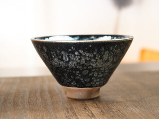 Front view of a Da Dou Li sky eye oil spot Jianzhan tea bowl on a wooden table, showing the wide flared rim, conical walls, small foot ring, and exterior silver oil-spot crystals.