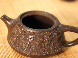 Close-up of carved calligraphy on the Guyun Shipiao Yixing teapot body, gritty antique bronze clay texture, bamboo mat