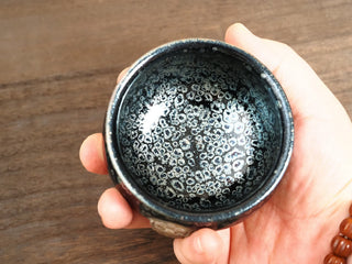 Handheld top-down view of a Rui Shou sky eye oil spot Jianzhan bowl, showing dense ring-shaped silver-blue crystals across the entire interior.