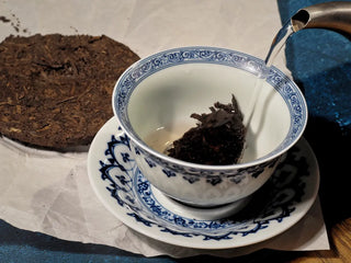 Hot water being poured over Yunnan Red Glow Ripe Pu-erh Tea Mini Cake – 100g in a white porcelain gaiwan, beginning to release its red-amber infusion.
