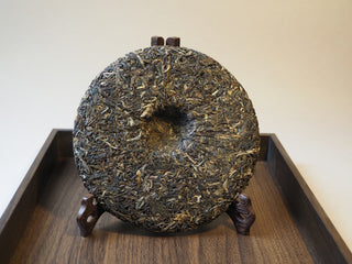 Back surface texture of the 2019 Tan Chun ancient tree raw puerh tea cake on a wooden stand.